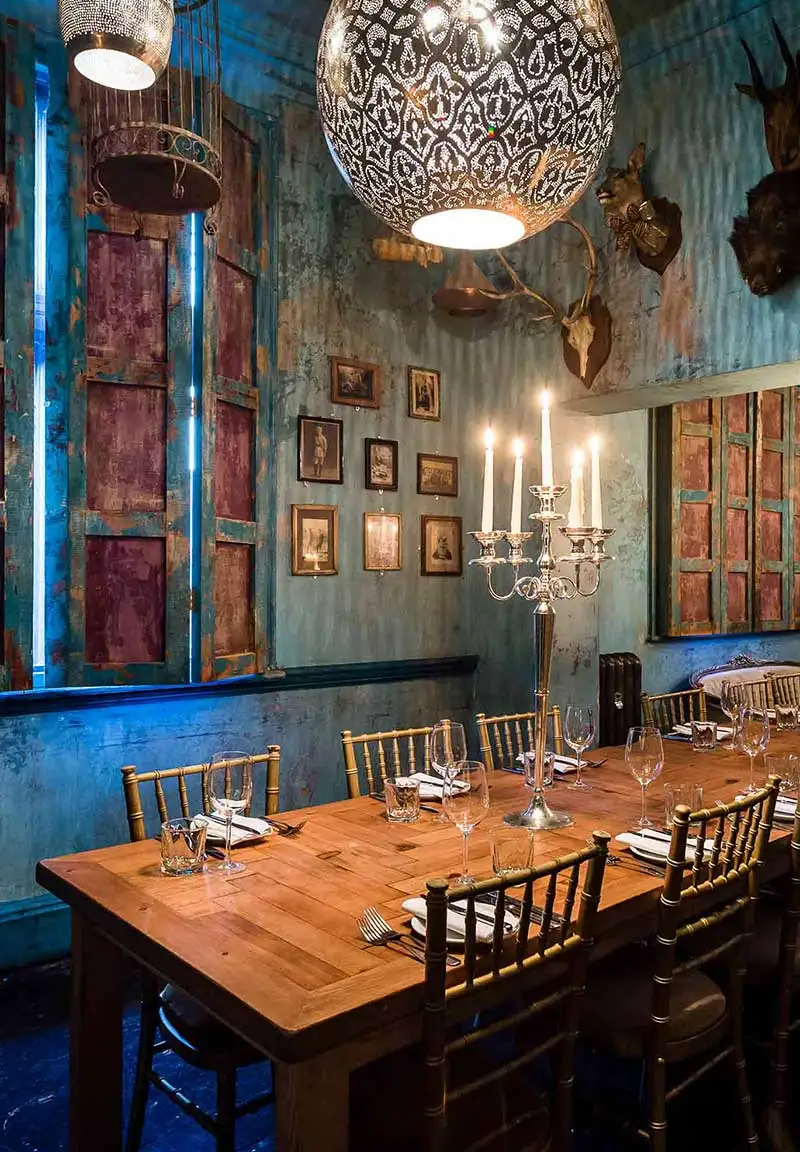 Rustic dining room with wooden table set with glassware, golden chairs, lit candle holder centerpiece, and blue distressed walls with framed pictures and mounted animal heads.