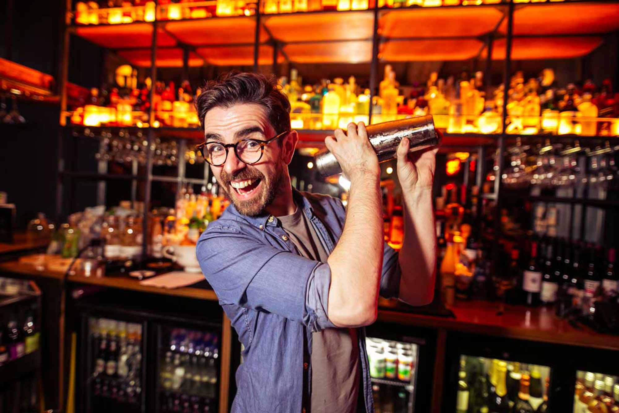 Smiling bartender with glasses shaking a cocktail shaker behind a bar with warm lighting and bottles in the background.