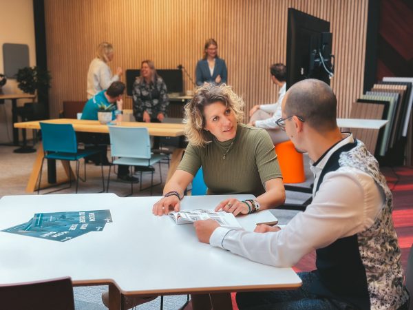 Twee mensen praten aan een tafel in een modern kantoor, met een groep mensen op de achtergrond.