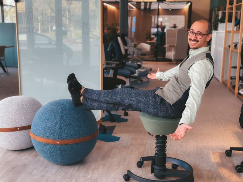 Man leunt achterover zittend op een bureaustoel met voeten op een blauwe bal in een kantoor.