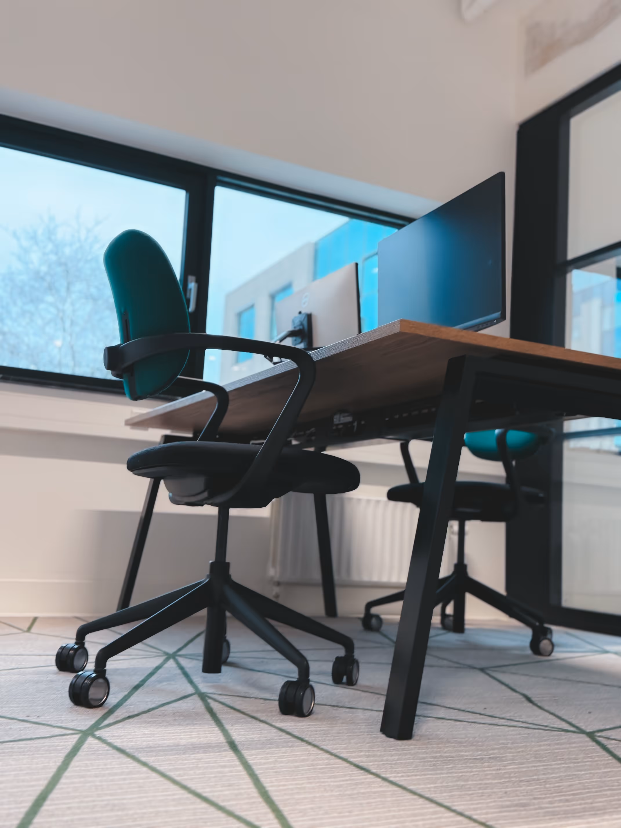 Twee bureaustoelen met groene rugleuningen bij een houten bureau met computerschermen in een kantoor.