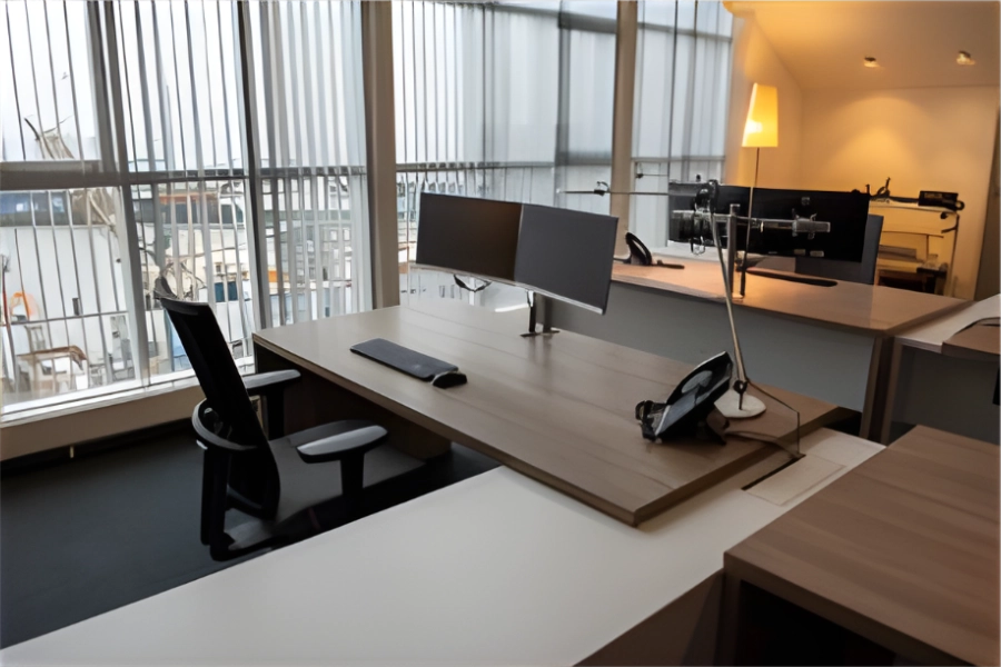 Modern kantoorruimte met bureaus, computerbeelden en ergonomische bureaustoel bij groot raam.