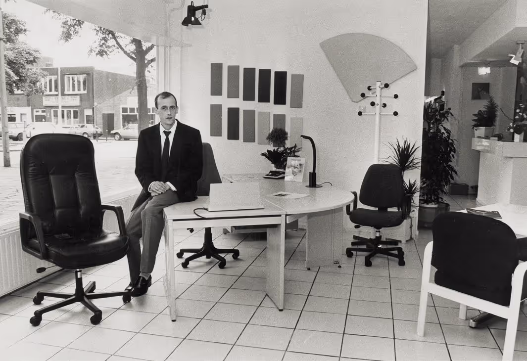Man in pak zit op een bureau in een kantoorruimte met bureaustoelen en planten.