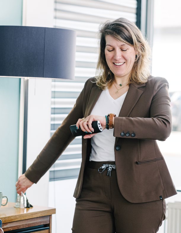 Vrouw in bruin pak belt een handbel in een lichte kamer met een zwarte lamp.