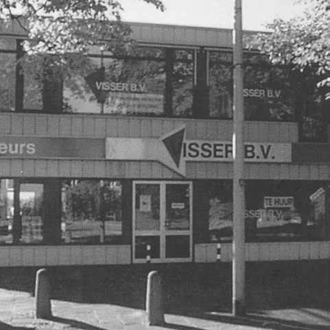 Zwart-witfoto van een bedrijfspand met het bord Visser B.V. en bomen erboven.
