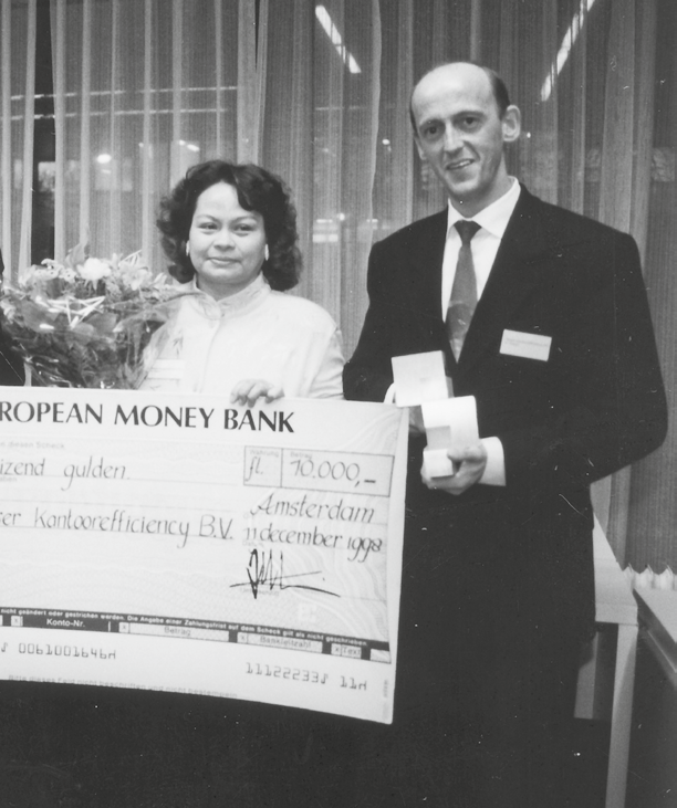 Twee personen houden een grote cheque van 70.000 gulden en een bos bloemen, zwart-wit foto.