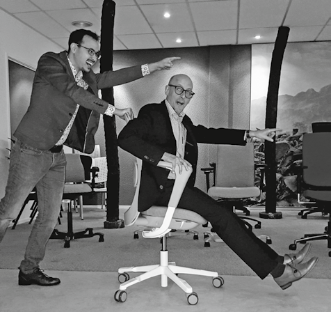 Twee mannen spelen lachend met een bureaustoel; een duwt terwijl de ander zit en wijst.