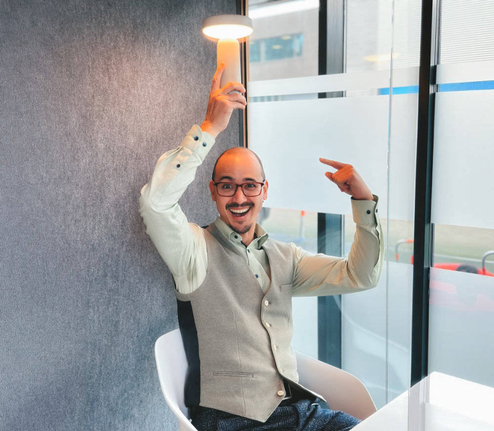 Man met bril en gilet zit en houdt een klein verlicht lampje boven zijn hoofd terwijl hij lacht.