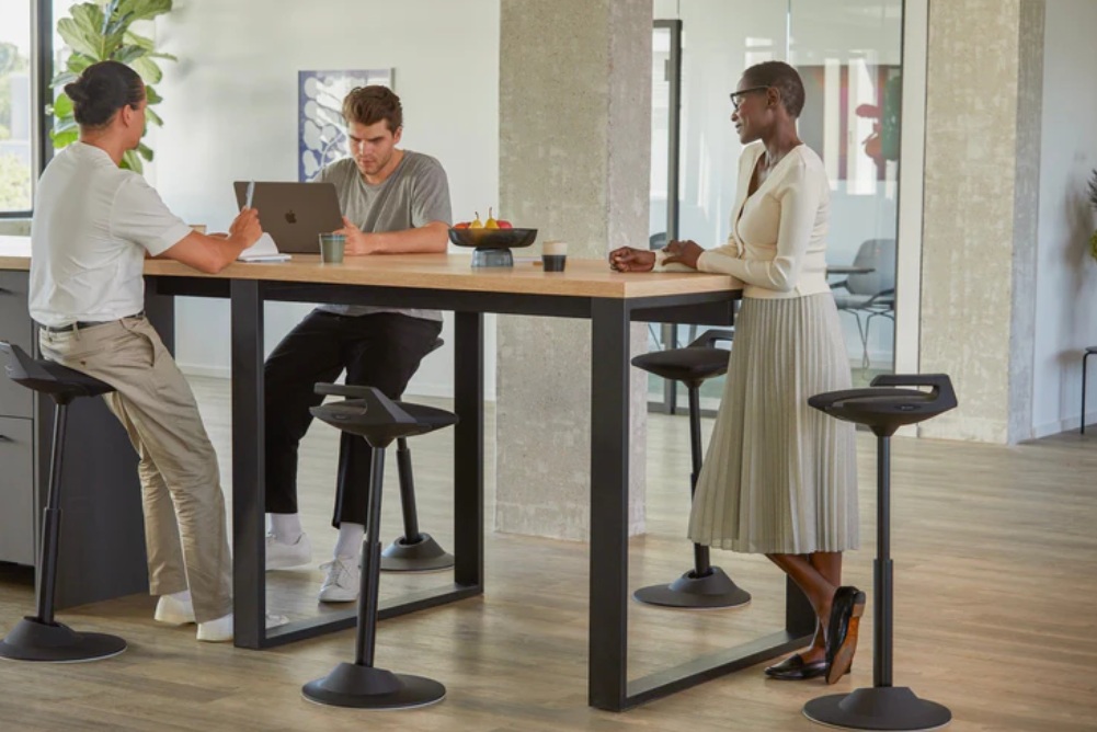 Drie mensen in een moderne kantoorruimte praten rond een hoge tafel met krukken.