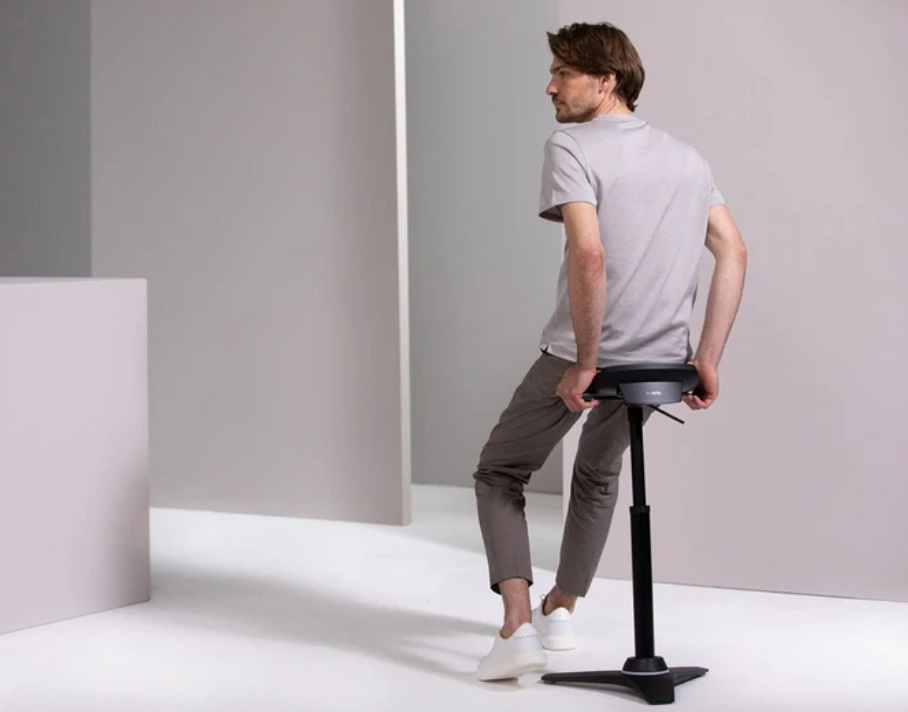 Man zit op een verstelbare ergonomische kruk met zwarte zitting in een neutrale ruimte.