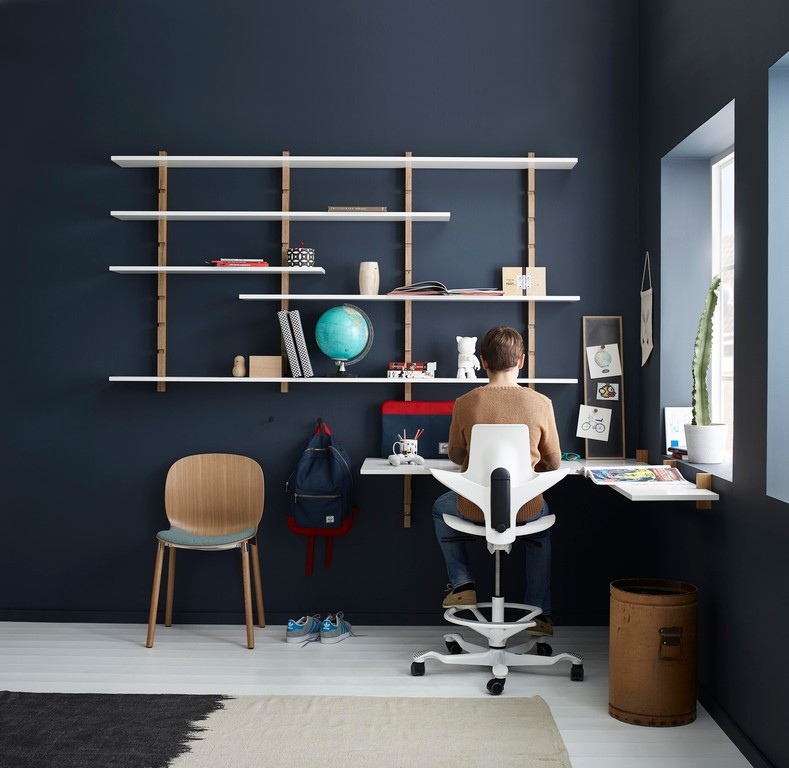 Persoon zittend op een ergonomische stoel aan een zwevend bureau met plank, wereldbol en raam.