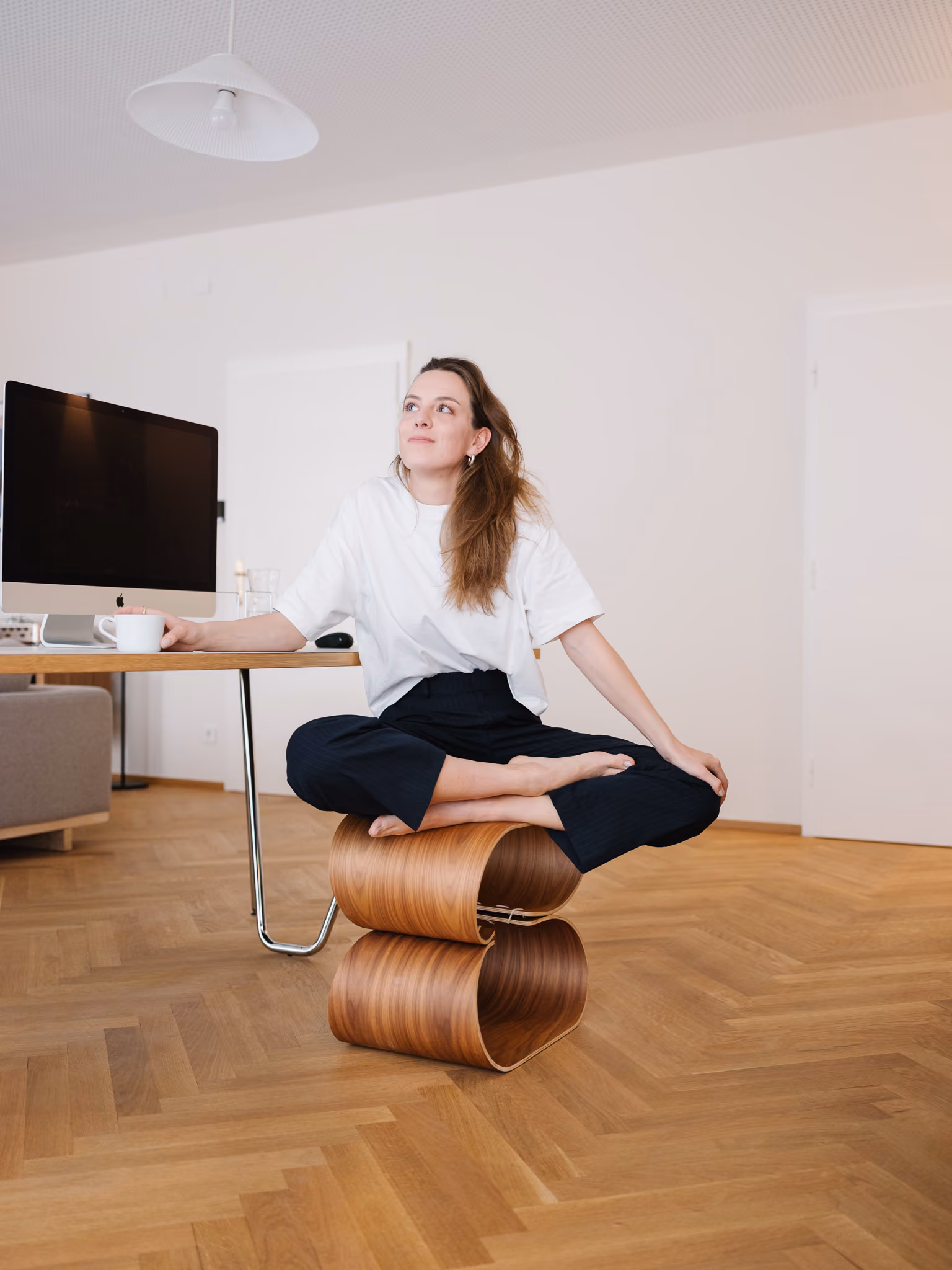 Vrouw in witte top zit gekruist op een golvende houten kruk bij een bureau met een computer.