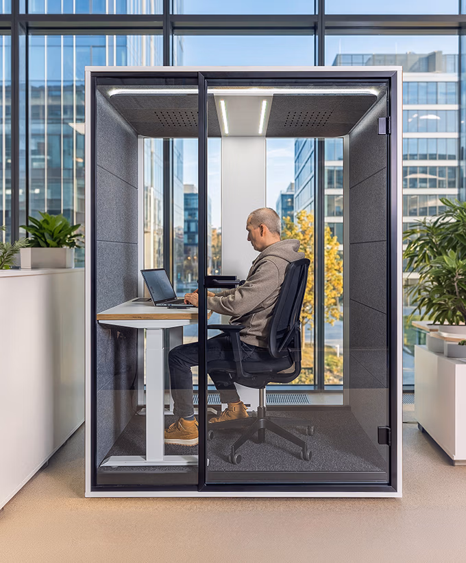 Man werkt op laptop in een afgesloten, geluidsdichte kantoorbox in een moderne kantoorruimte.