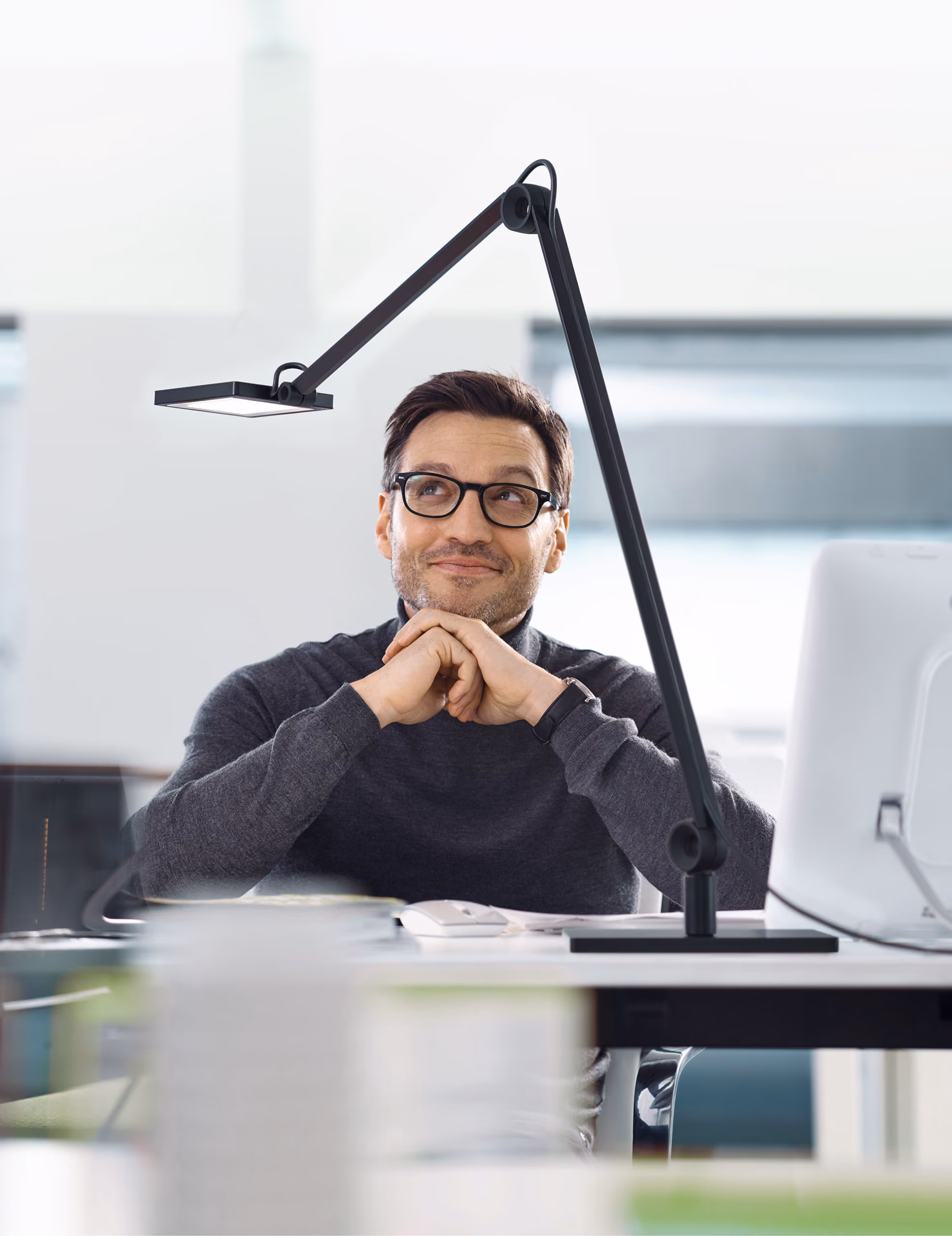Man met bril en grijze trui kijkt dromerig bij een bureau met bureaulamp en computerscherm.