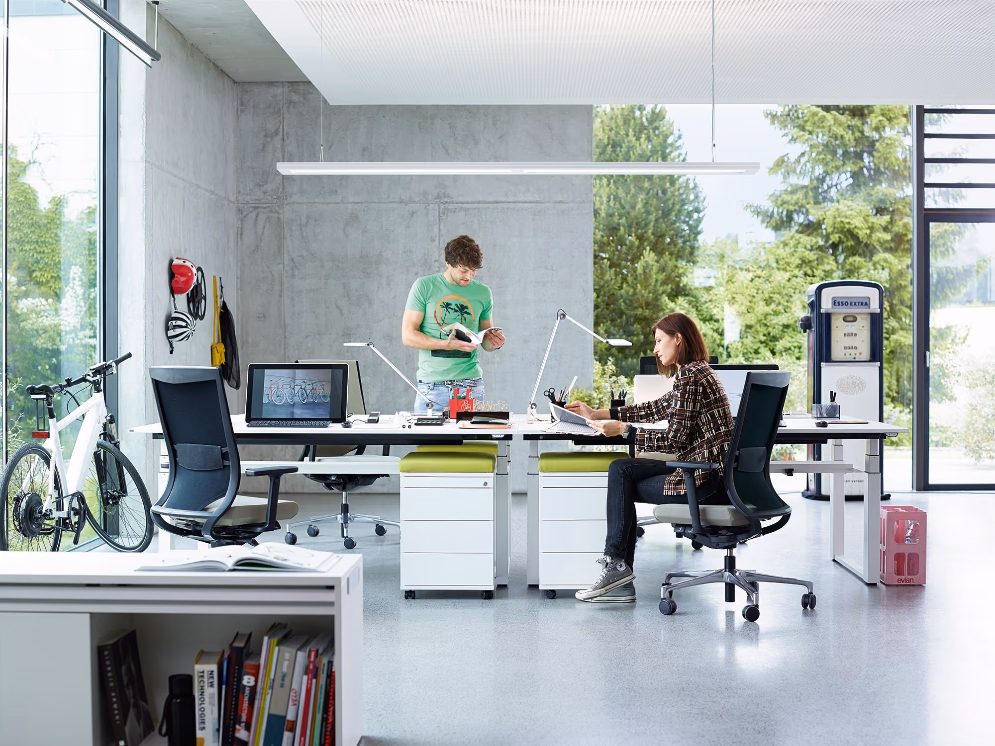 Moderne kantoorruimte met grote ramen en zicht op groen buiten, met twee mensen aan het werk: een man staat bij een bureau en leest een boek, een vrouw zit en werkt met documenten, rondom bureaus staan bureaustoelen, een fiets, een vintage brandstofpomp en boekenplanken zijn zichtbaar.