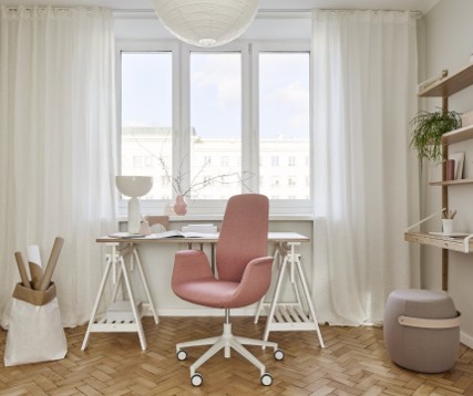 Een modern thuiskantoor met een roze ergonomische bureaustoel op wieltjes voor een wit bureau met houten poten, een witte lamp, en een roze vaas. Grote ramen met witte gordijnen laten veel licht binnen. Rechts staan planken met planten en boeken, en een grijze poef staat op de houten visgraatvloer.