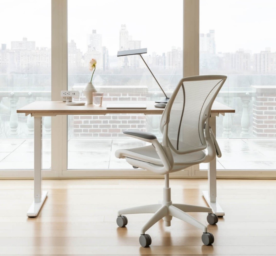 Minimalistisch bureaumeubilair met een lichte, moderne bureaustoel met gaasrugleuning en een houten verstelbaar bureau geplaatst voor een groot raam met uitzicht op de stad.