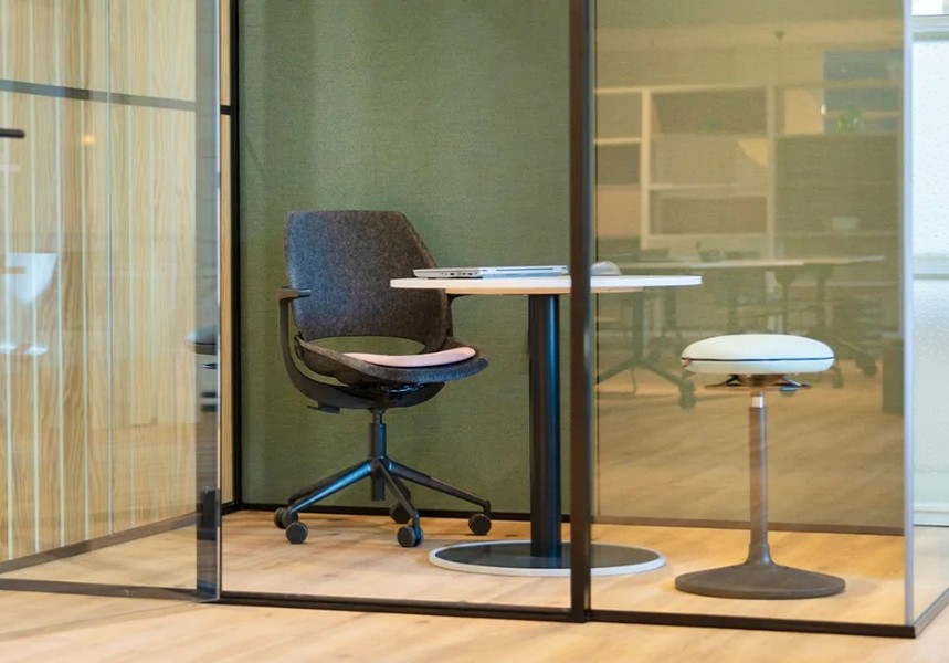Een vergaderruimte met glazen wanden, ingericht met een zwarte bureaustoel met wieltjes, een ronde tafel met een laptop en een witte kruk zonder rugleuning.