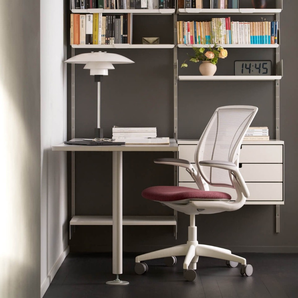 Moderne werkruimte met witte bureaustoel met een rood zitting, een minimalistisch wit bureau met boeken en een lamp erop, en een wandplanksysteem met boeken, een digitale klok die 14:45 toont, en een vaas met bloemen.