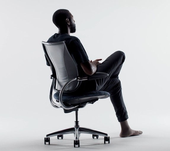 Een man zit ontspannen achterover leunend in een ergonomische bureaustoel met een mesh rugleuning en metalen onderstel op wieltjes tegen een witte achtergrond.