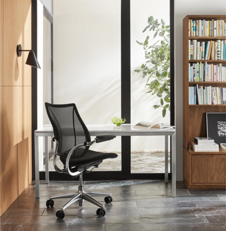 Moderne kantoorruimte met een zwarte ergonomische bureaustoel met gaasrugleuning, een glazen bureau met metalen poten, een boekenkast vol boeken en een groene plant, alles tegen een lichte muur met een groot raam.