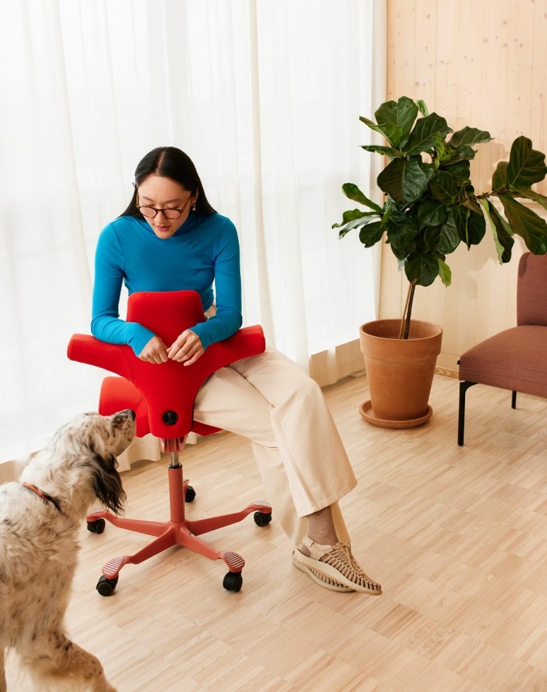 Vrouw in een blauw shirt en beige broek zit omgekeerd op een rode bureaustoel terwijl ze naar een witte hond met zwarte vlekken kijkt in een lichte kamer met houten vloer en een grote groene plant in een pot.