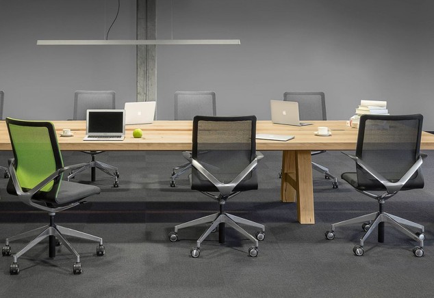 Moderne vergadertafel van licht hout met zes zwarte bureaustoelen met mesh-achterkant, laptops, boeken, kopjes en een groene appel erop, in een minimalistisch kantoor met grijze muren en hanglamp.