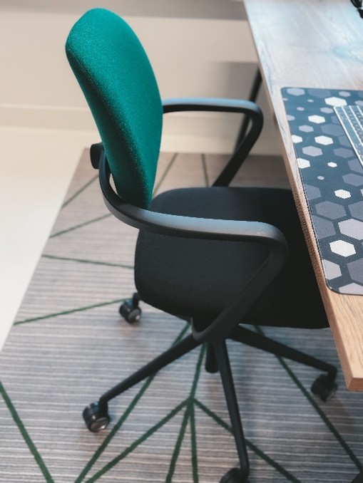 Bureaustoel met groene rugleuning en zwarte zitting, met zwarte armleuningen en op zwarte wieltjes, geplaatst naast een houten bureau met een zwart witte hexagon-gepatteerde muismat.