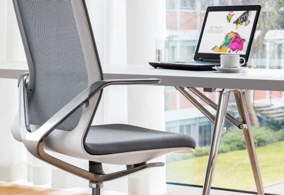 Moderne ergonomische bureaustoel met grijze mesh zitting en rugleuning naast een tafel bij een raam, met een opengeslagen laptop en een koffiekopje erop.