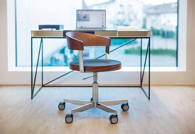 Moderne bureaustoel van hout en metaal met wielen, geplaatst voor een minimalistisch bureau met een laptop, notitieboek en boeken, bij een groot raam.