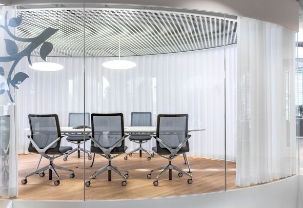 Moderne vergaderruimte met glazen wanden, zes grijze bureaustoelen op wielen rondom een ovaal witte tafel, houten vloer, witte gordijnen en twee hanglampen aan het plafond.