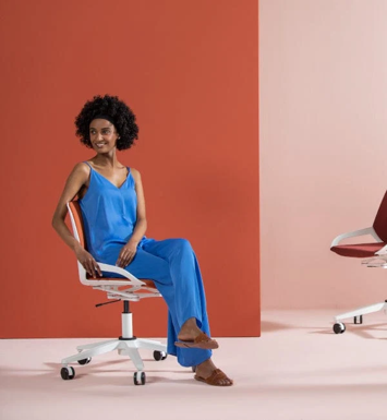 Vrouw zittend op een witte bureaustoel met wielen, gekleed in een blauwe jumpsuit en bruine sandalen, tegen een roze en oranje achtergrond.