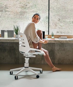 Een vrouw zittend op een witte bureaustoel met wieltjes in een lichte kamer met betonnen vensterbank en enkele planten, kijkend en glimlachend naar rechts.