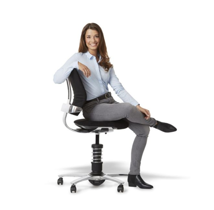 Een vrouw met lang bruin haar draagt een lichtblauw overhemd, grijze broek en zwarte laarzen, en zit ontspannen met gekruiste benen op een zwart ergonomische bureaustoel met zilveren onderstel en wieltjes tegen een witte achtergrond.