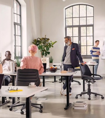 Een moderne kantoorruimte met vier mensen: een man met grijs haar en baard in een donker colbert leunt tegen een stoel, een vrouw met roze haar zit aan een bureau, een man met een gestreept shirt staat bij een raam en een andere man zit aan een bureau. Grote ramen en een groene plant zijn ook zichtbaar.