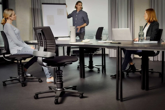 Drie volwassenen in een kantoorruimte tijdens een bespreking; een man staat bij een flip-over met bescheiden tekeningen, terwijl twee vrouwen op bureaustoelen luisteren, een met laptop en een met armen gekruist.