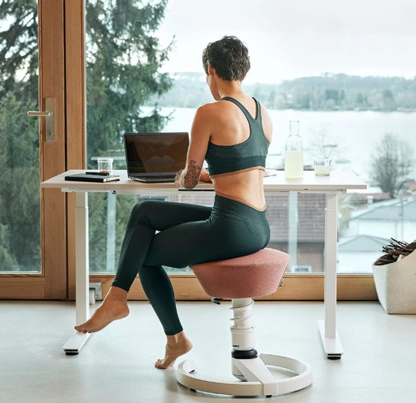 Een vrouw in sportkleding zit op een ergonomische stoel aan een bureau met een laptop, met uitzicht op bomen en een meer buiten een groot raam.