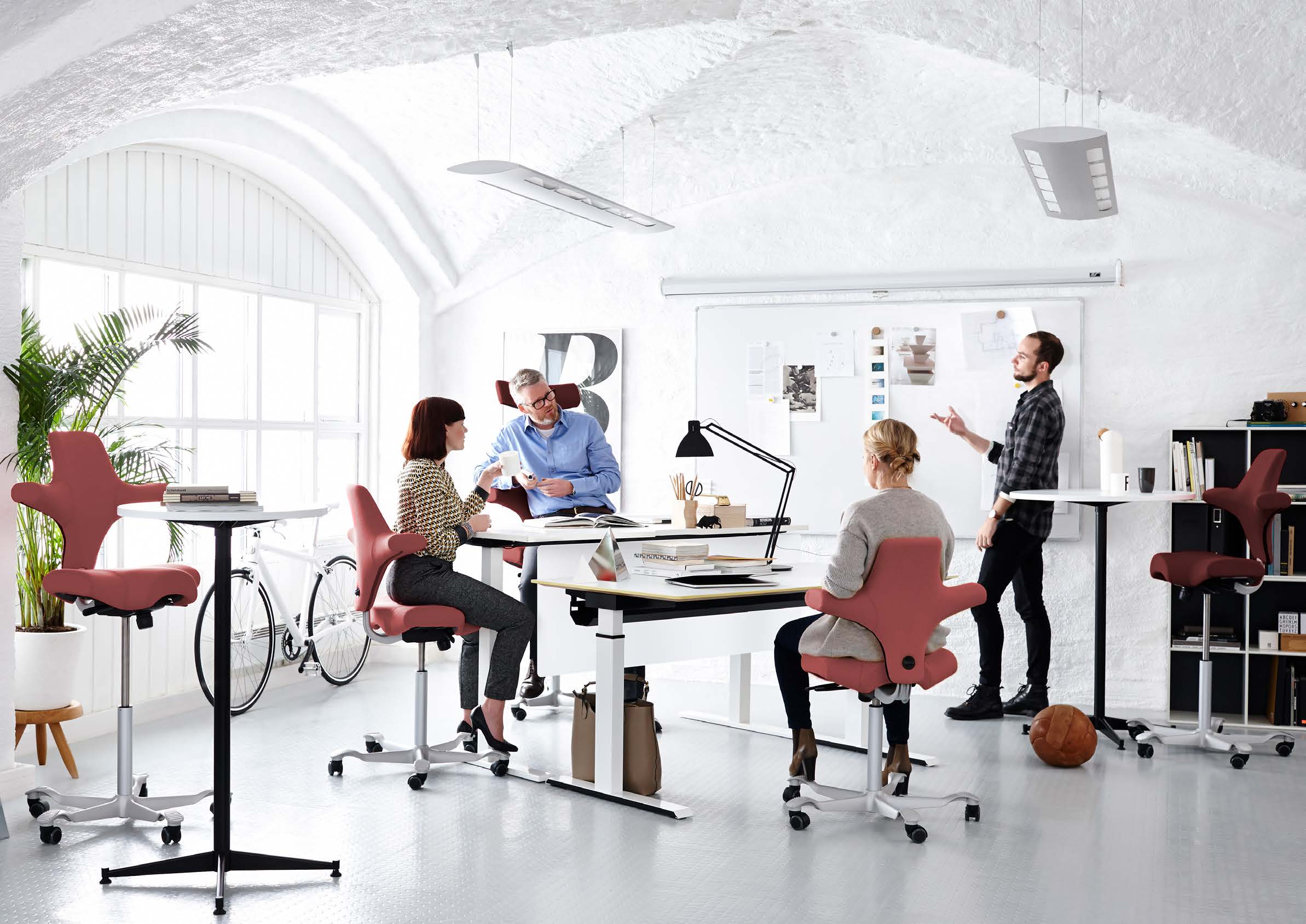 Vier mensen in een lichte, moderne kantoorruimte met witte gewelfde muren en grote vensters; drie zitten in roze ergonomische bureaustoelen rond twee witte tafels, een man staat voor een whiteboard met papieren er op, terwijl ze lijken te overleggen.