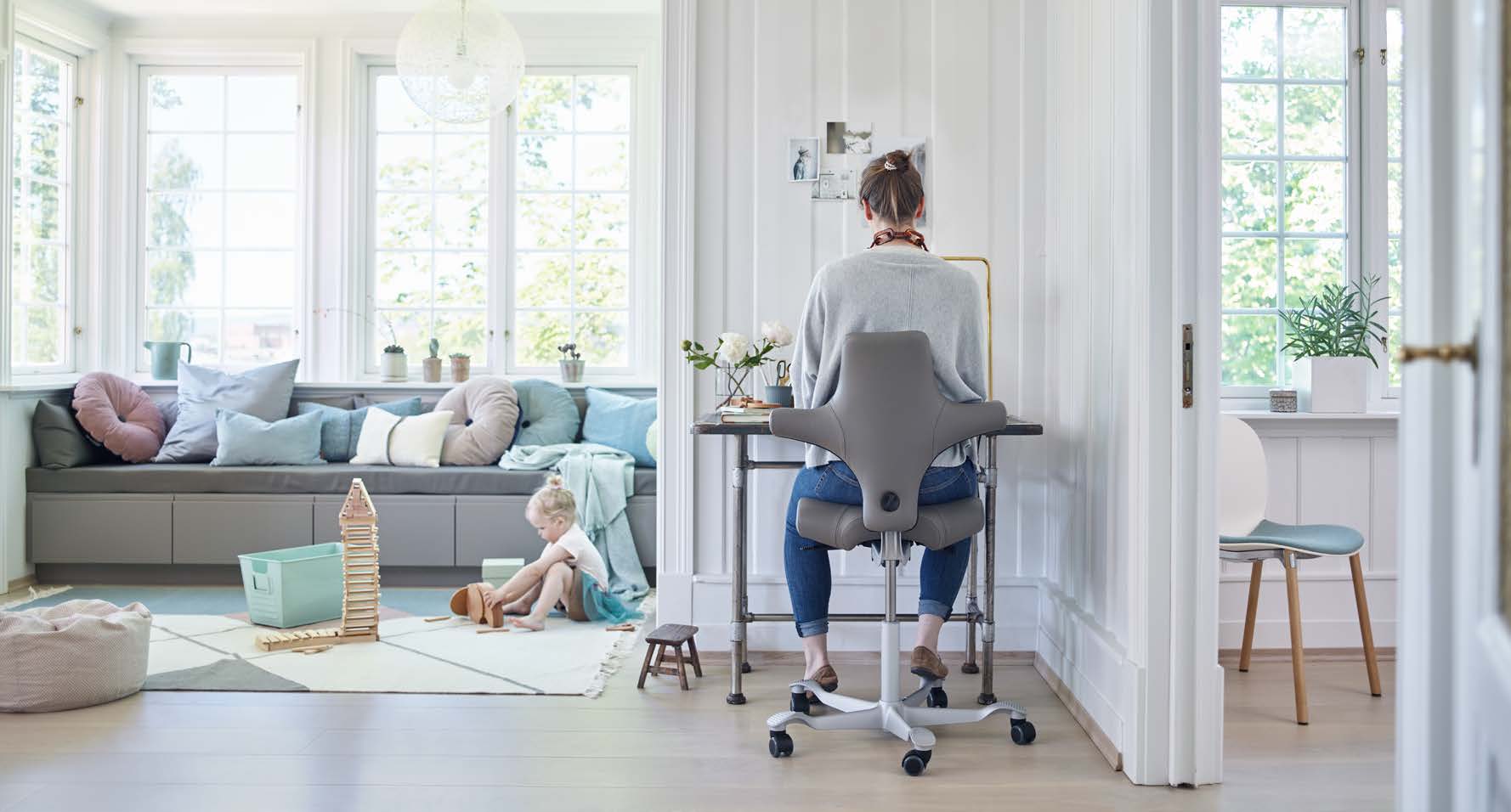 Vrouw zittend aan een bureau in een helder thuiskantoor, met een jong kind spelend op een vloerkleed bij een raam met veel kussens.
