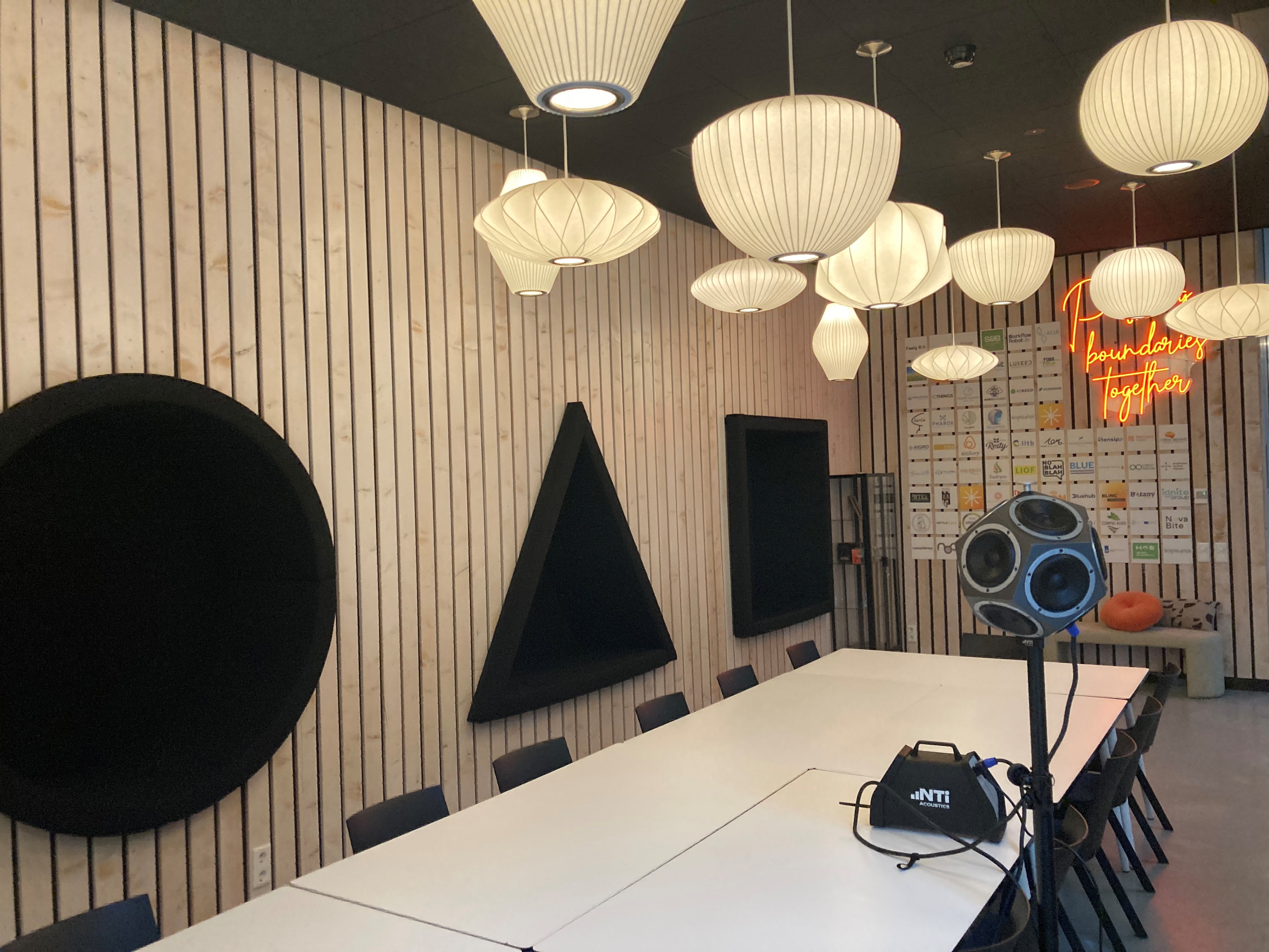 Vergaderruimte met een lange witte tafel omringd door zwarte stoelen, unieke geluidsabsorberende wandpanelen in cirkel, driehoek en vierkant, verschillende hanglampen en een meetinstrument op een statief met het logo NTi Acoustics, neonbord met tekst 'No boundaries together'.