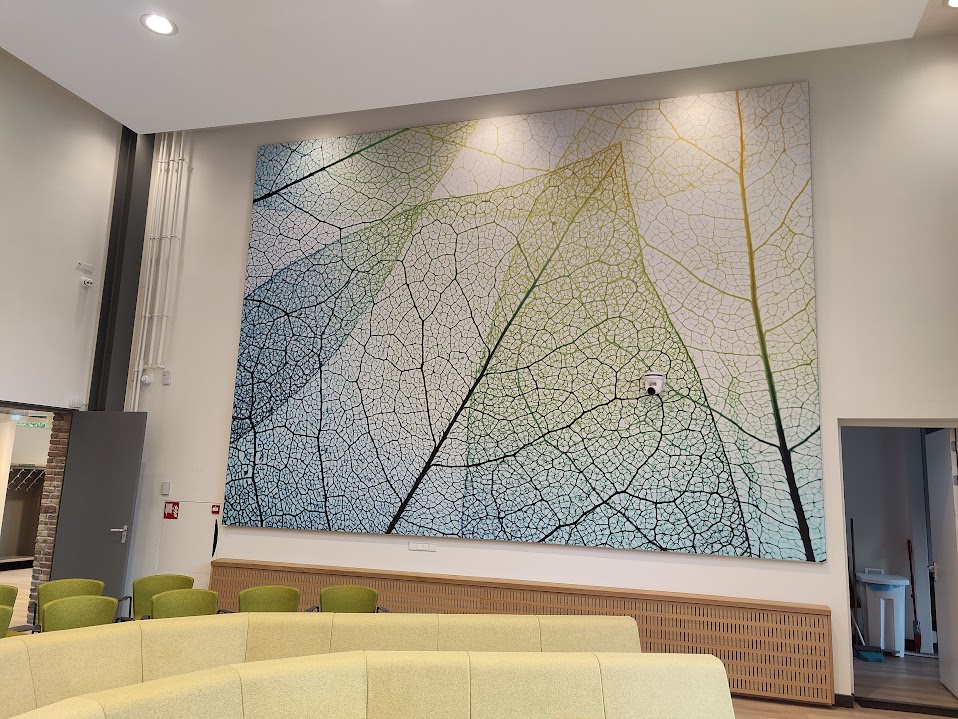 Een modern interieur met een grote, kleurrijke muurschildering van verstrengelde bladeren en nerven, omgeven door lichte muren, groene stoelen en een gebogen bank.