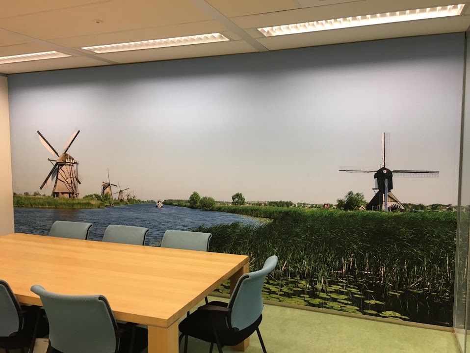 Vergaderruimte met een houten tafel en zes lichte stoelen, een grote muurfoto van een rivierlandschap met riet en traditionele Nederlandse molens onder een heldere blauwe lucht.