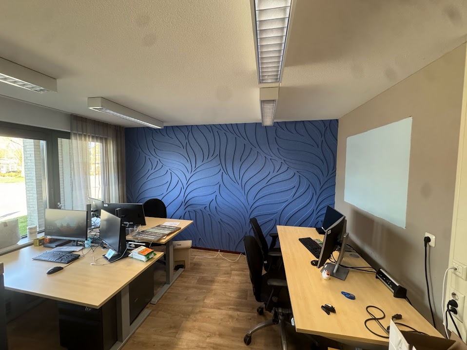 Kantoorruimte met twee houten bureaus, elk voorzien van computerschermen, toetsenborden, muizen en bureaustoelen, met een blauwe wand met golvend patroon aan de achterzijde en een blanco whiteboard aan de rechtermuur.