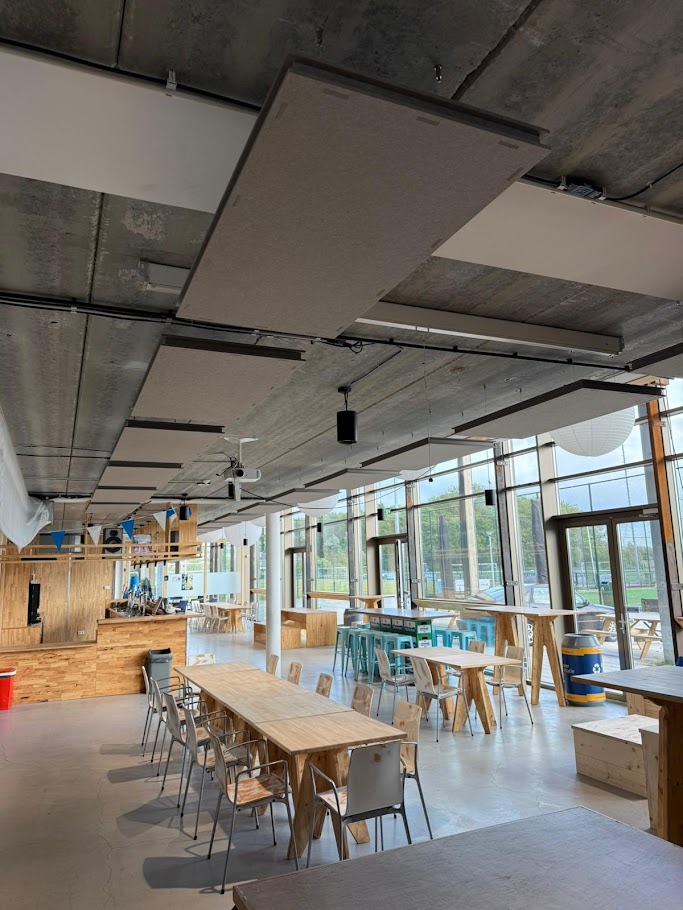 Een ruime, lichte eetzaal met houten tafels en stoelen, metalen krukken aan een lange hoge tafel, grote ramen langs de rechterkant, een betonnen plafond met geluidsabsorberende panelen en industriële verlichting.