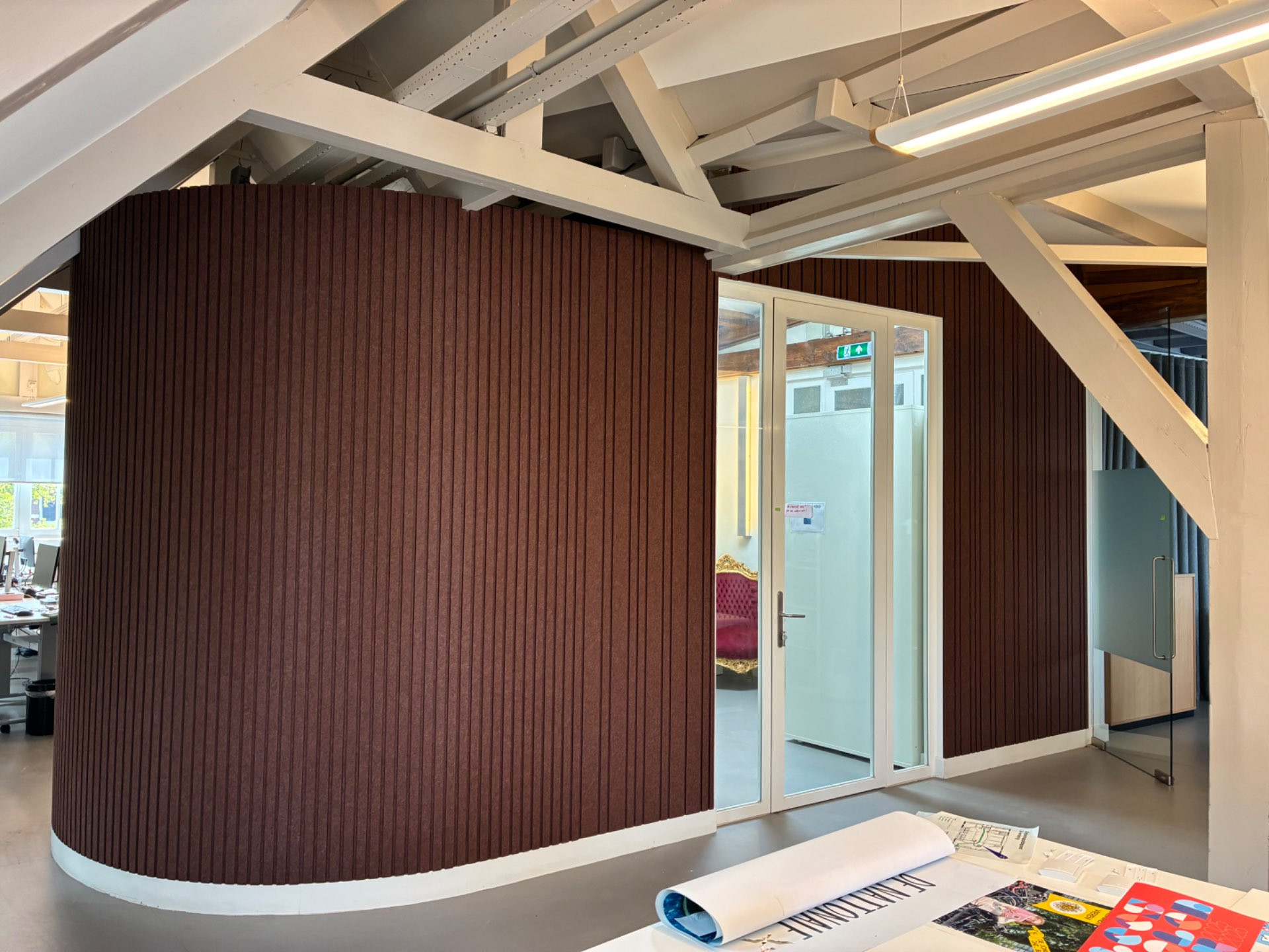 Moderne kantoorruimte met een gebogen muur bekleed met verticale bruine panelen en een glazen deur, witte balken aan het plafond en een tafel met opgerolde documenten en kleurrijke tijdschriften erop.