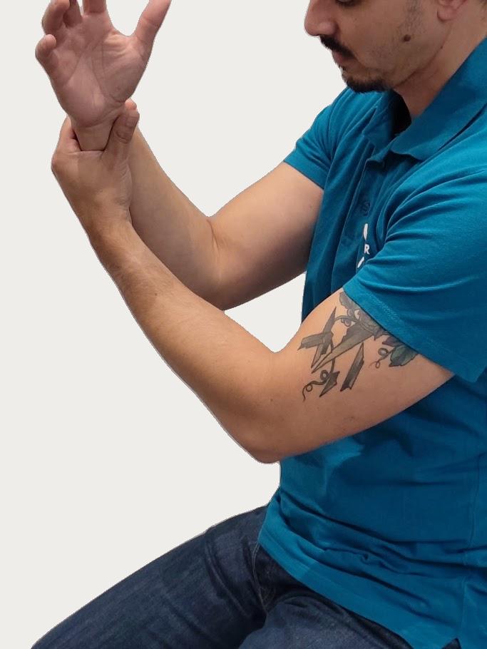 Man met tatoeage op de rechterbovenarm die zijn linkerpols vasthoudt en zijn elleboog strekt, zittend en in een blauw poloshirt.