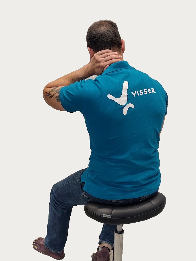Man zittend op een kruk met zijn rug naar de camera gekeerd, draagt een blauw poloshirt met het woord Visser en een logo, hij houdt zijn nek met een hand vast alsof hij nekpijn heeft.