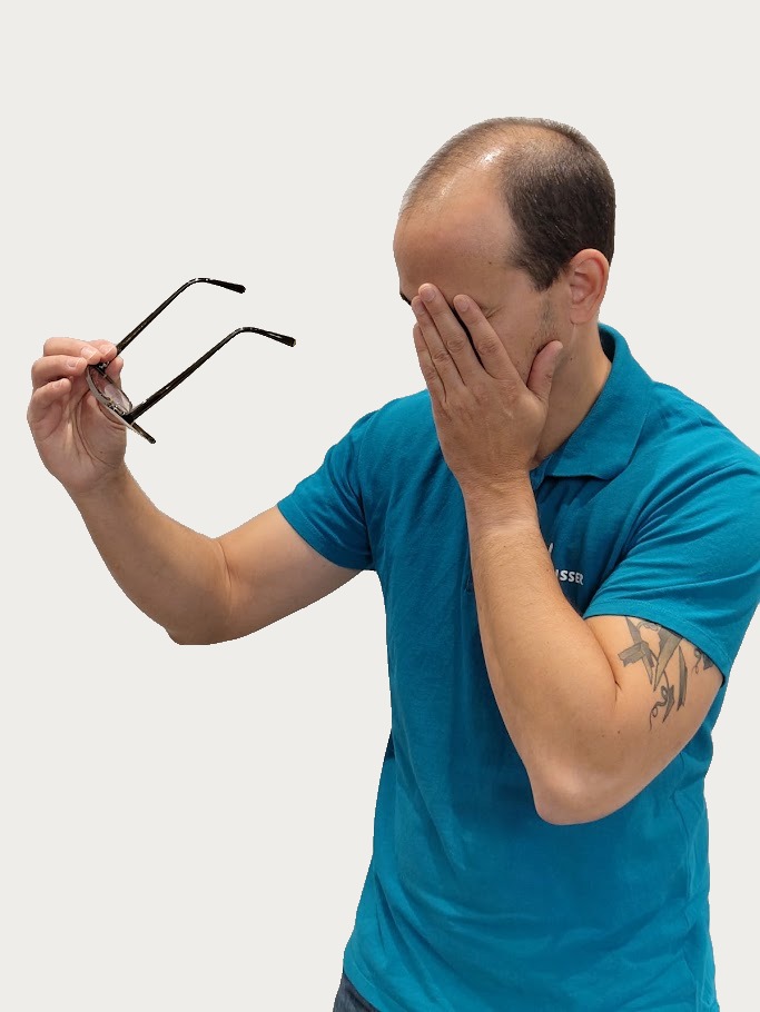 Man met blauwe polo die zijn gezicht met één hand bedekt en in de andere hand een bril vasthoudt, zichtbaar ongemakkelijk met dichtgeknepen ogen.