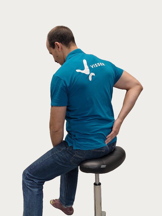 Man zittend op een kruk met een blauwe polo met het woord 'VISSER' op de rug, die zijn onderrug vasthoudt alsof hij pijn heeft.
