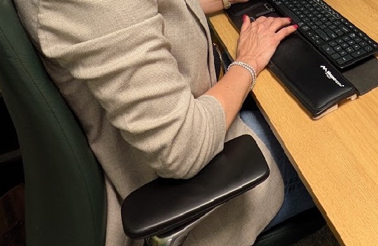 Vrouw zittend aan een bureau met haar armen op een zwarte, ergonomische armsteun terwijl ze typt op een toetsenbord.